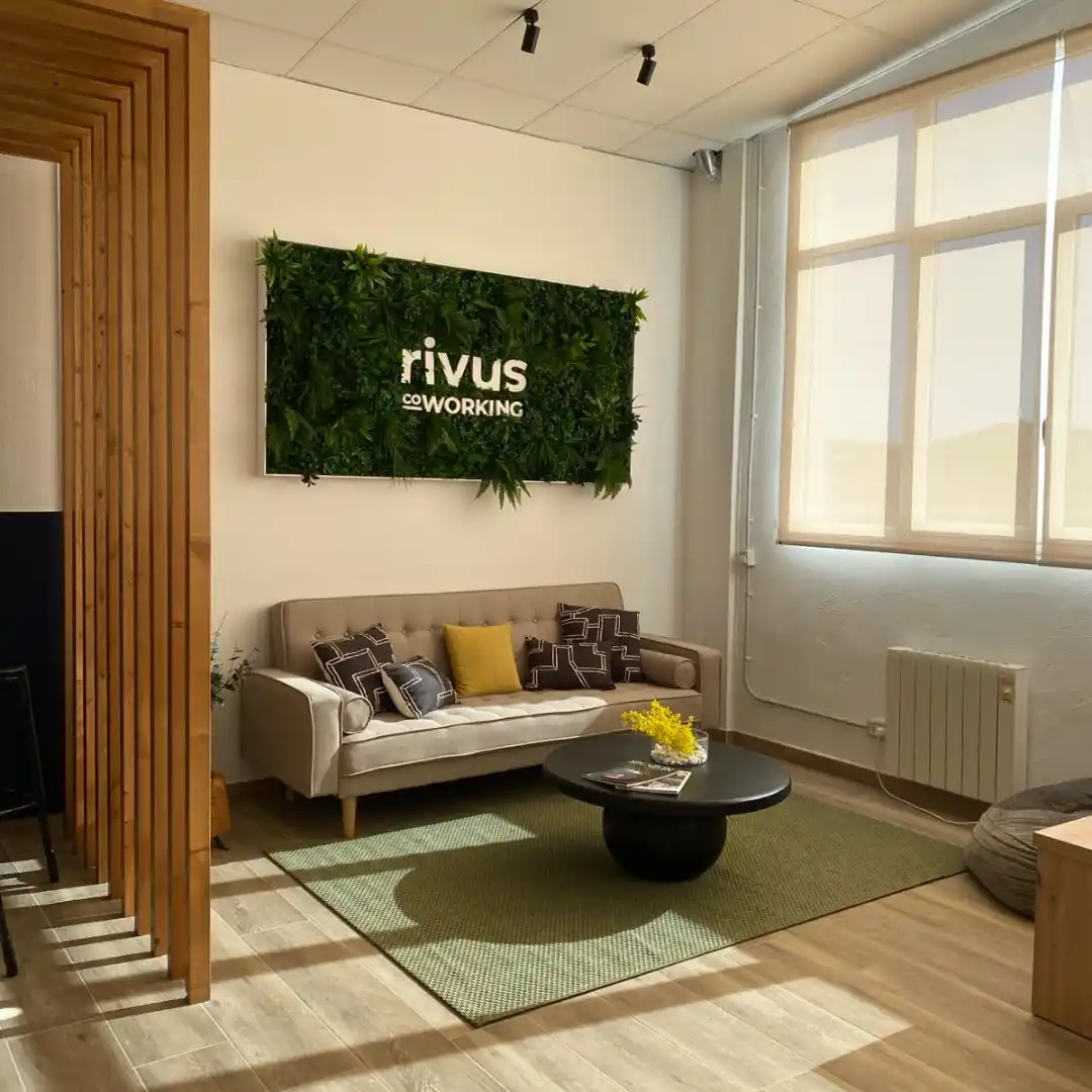 Área de descanso en el Coworking Rivus con un sofá cómodo, mesa baja y decoración verde en la pared.