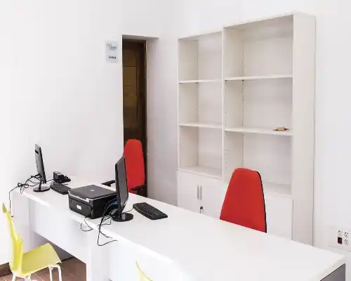 Oficina en el Centro Pedrola con mesa larga blanca, equipos de computación, sillas rojas y mueble de guardado blanco.