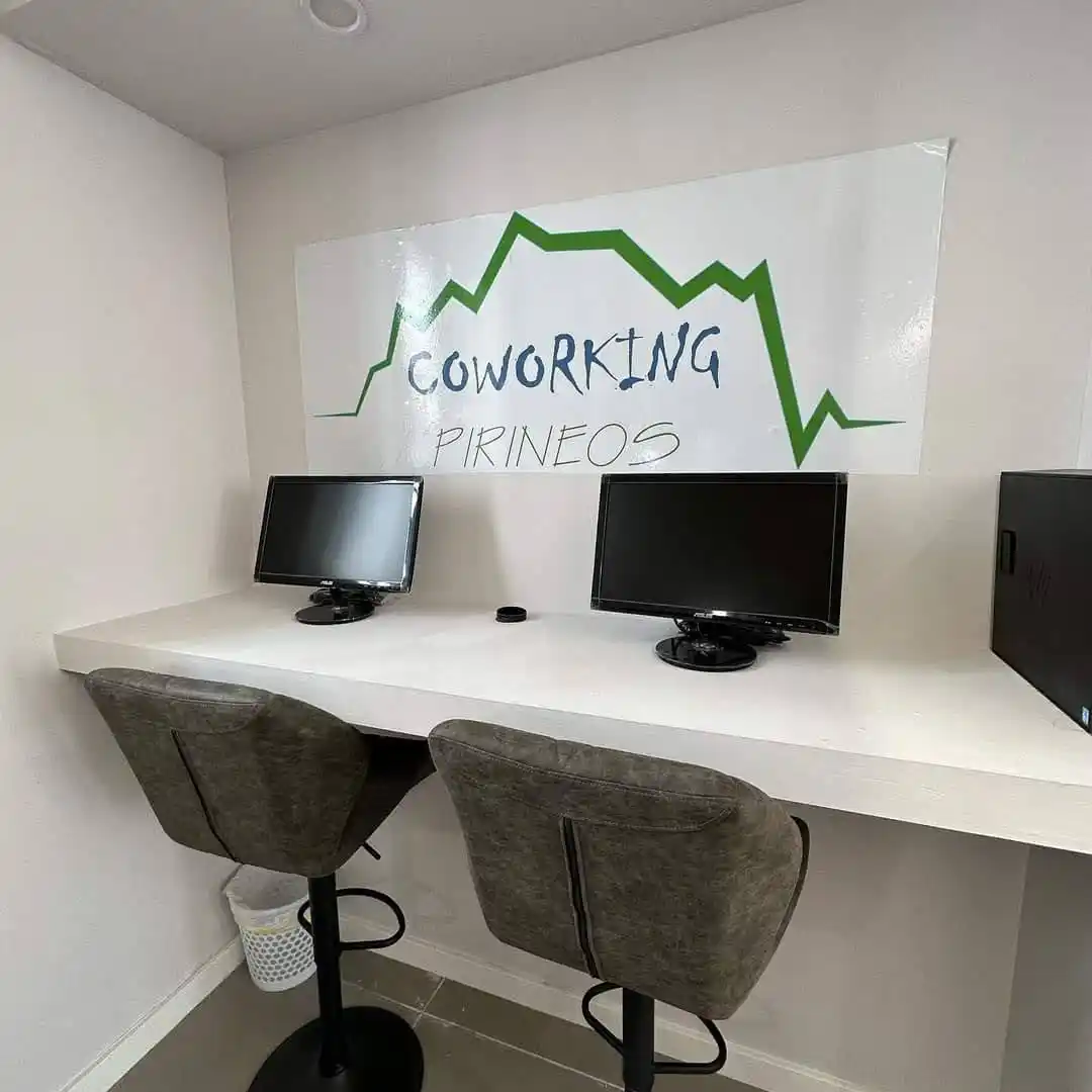Espacio de trabajo en el Centro Pirineos con una mesada equipada con computadoras, sillas negras y un mural en la pared que muestra el logo del centro.
