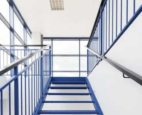 Escalera metálica en el centro Graus con pasamanos azules y grandes ventanales.