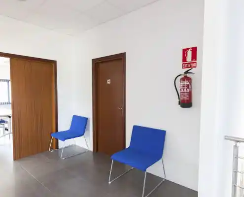 Pasillo en el centro Mequinenza con sillas azules y extintor de incendios.