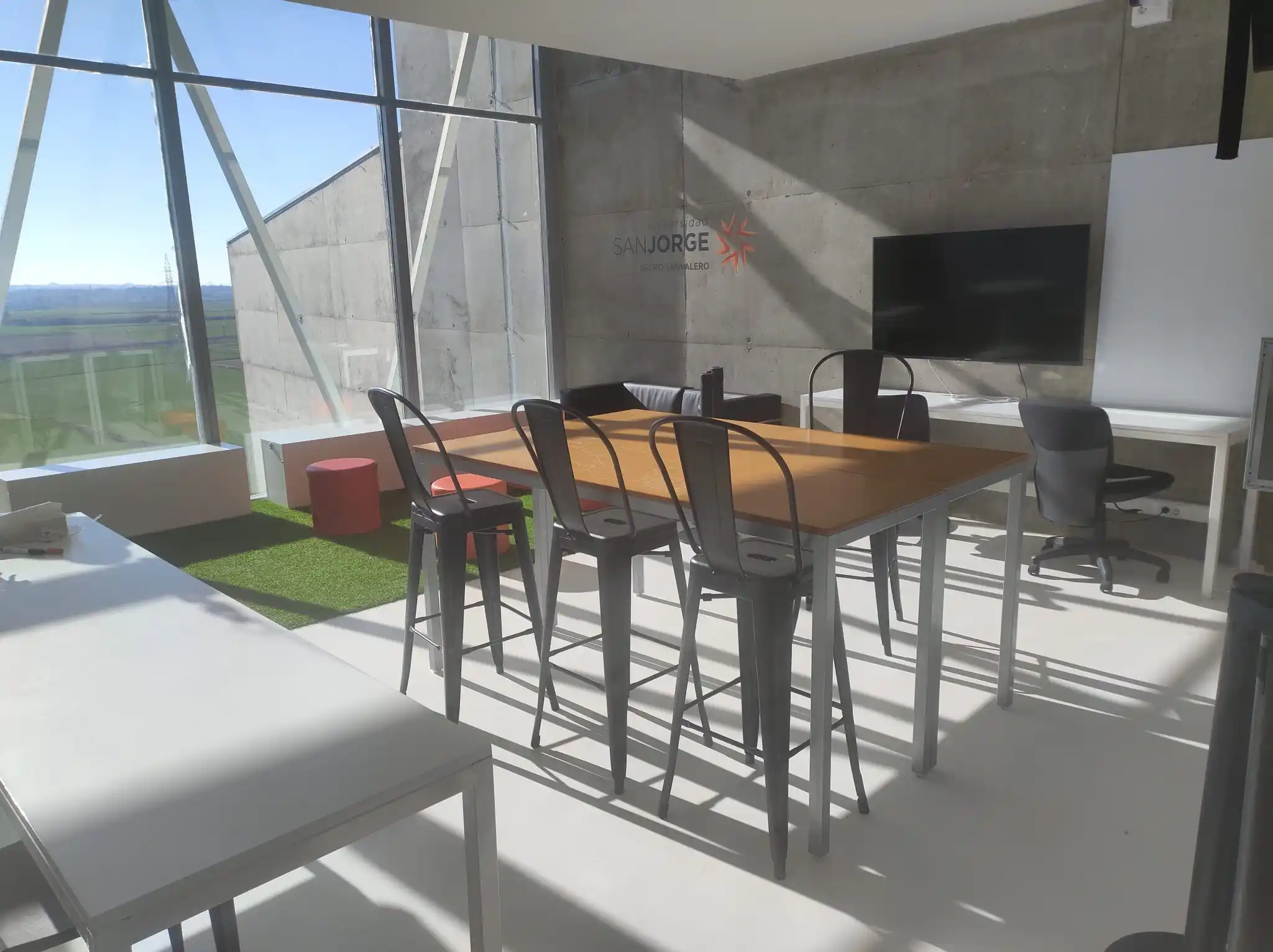 Oficina luminosa en El Cubo de las Ideas, con un amplio ventanal, mesas y sillas de diseño moderno, y una pantalla montada en la pared.