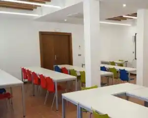 Salón del Centro Pedrola con paredes blancas, puerta de madera y mesas largas con sillas de colores variados.