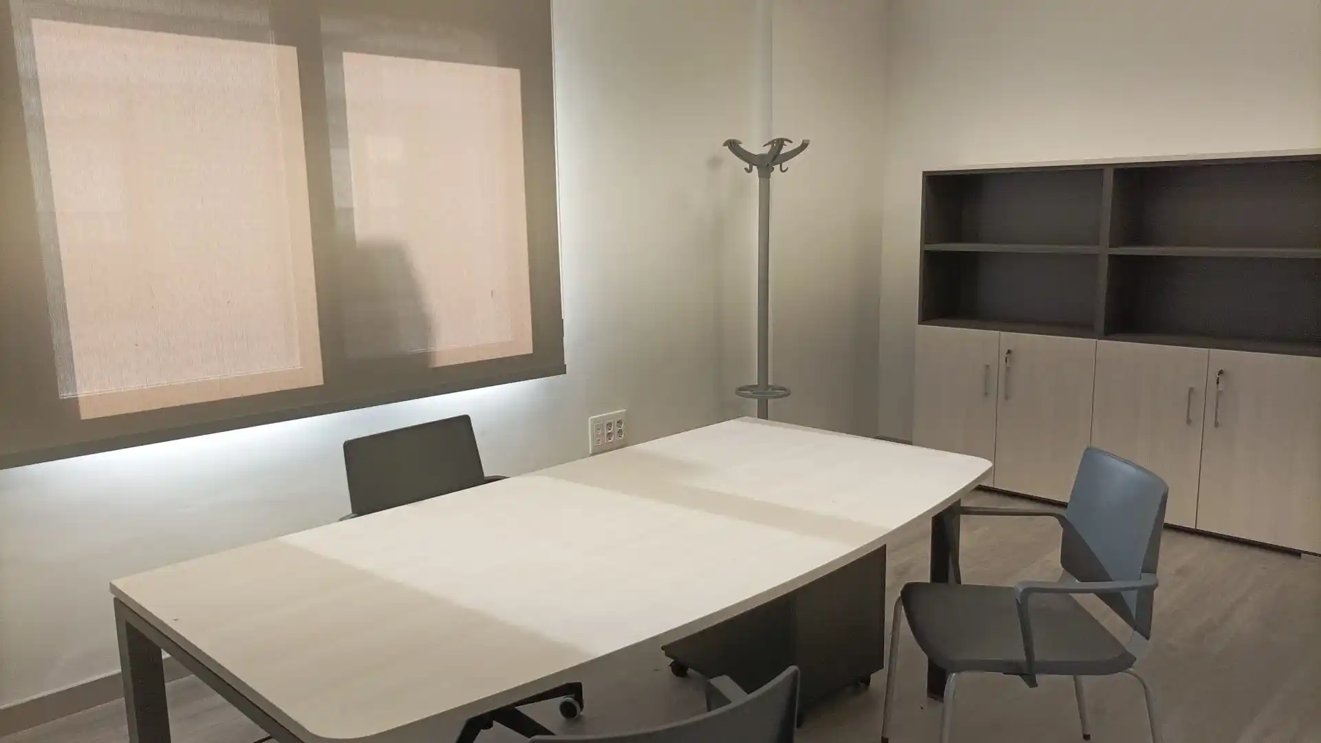 Oficina con mesa grande blanca, sillas negras, perchero y ventana.