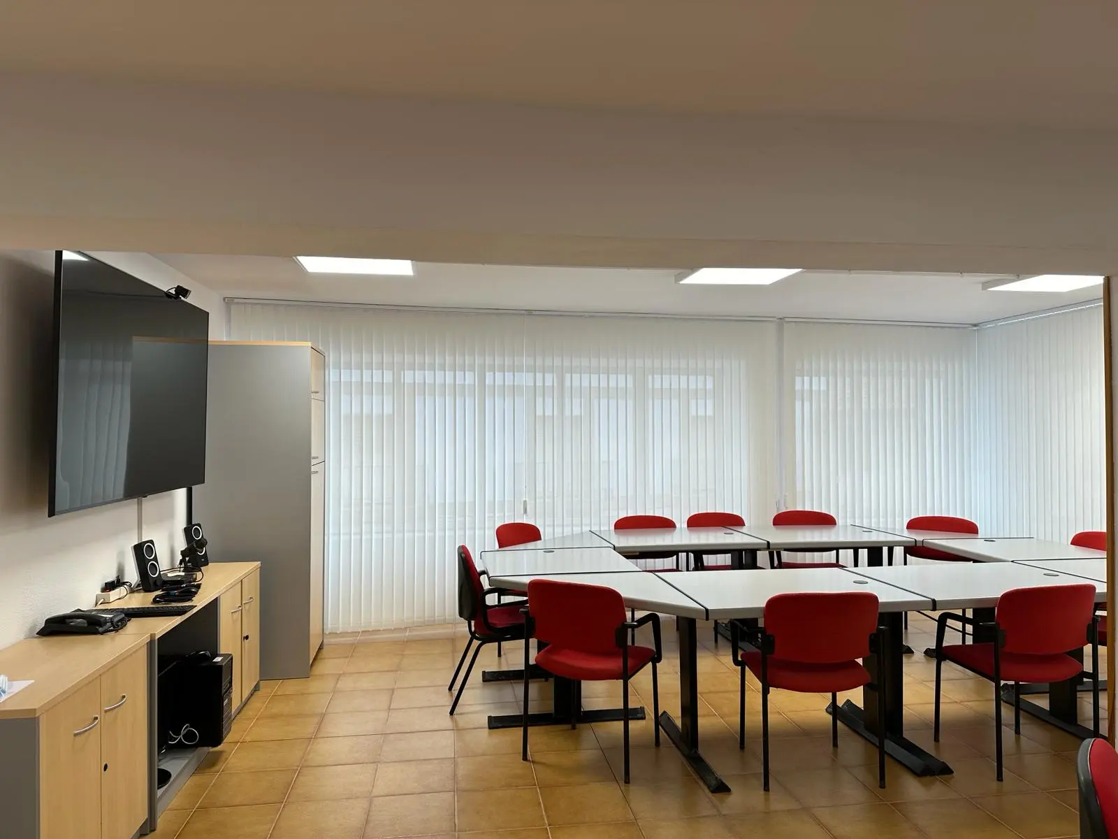 Sala de reuniones del Centro Ceder Monegros con mesas blancas y sillas rojas, equipada con pantalla y equipo audiovisual.